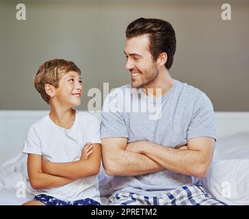 Following in his fathers footsteps. a cheerful father and son seated on a bed with arms folded while looking at the camera at home in the morning. Stock Photo