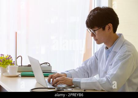 Young doctor physician wearing white uniform and using laptop in hospital office. Male doctor in eyeglasses typing on his computer at desk Stock Photo