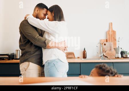 Cosy couple hugging, romance and affection in a modern kitchen with small child looking at them. Loving, young and parents, mom and dad embrace, excited by the future of their growing family Stock Photo