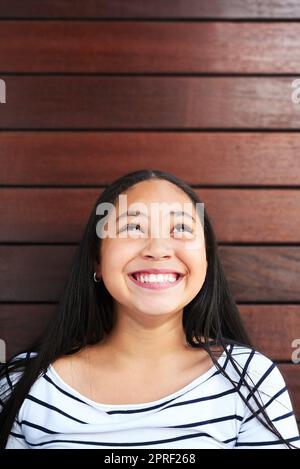 She already knows the secret to life. Keep looking up. a happy young girl posing against a wooden background and looking up. Stock Photo