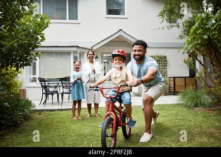 Boy on bicycle learning with proud dad and happy family in their home garden outdoors. Smiling father teaching fun skill, helping and supporting his excited young son to ride, cycle and pedal a bike Stock Photo