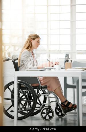Professional, disabled business woman in wheelchair reading documents, writing or making notes on office desk sitting by laptop. Female entrepreneur with disability doing contract paperwork with pen. Stock Photo