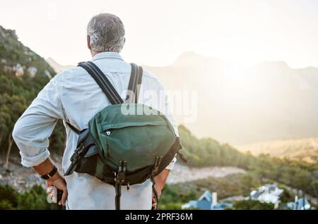 . Rear view of exploring, active and adventure senior man standing with a backpack after a hike, enjoying the landscape and forest nature. Male hiker looking at nature environment sunrise view. Stock Photo