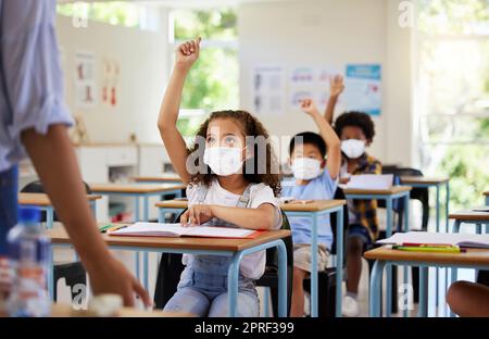 Covid, education and learning of eager, smart and clever children raising hands and wearing masks in a classroom. Teacher or educator asking questions for knowledge test at kids school in a pandemic. Stock Photo