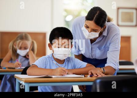 Covid, education and learning with a teacher wearing a mask and helping a male student in class during school. Young boy studying in a classroom with help from an educator while sitting at his desk. Stock Photo