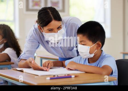 Covid, students and education teacher helping learning children in school classroom to answer math question. Woman with face mask teaching asian boy in lesson with diverse class of kids or classmates Stock Photo