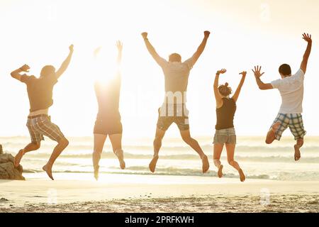 Reach for the sky. Rearview shot of a group of unidentifiable friends jumping together on the beach at sunset. Stock Photo