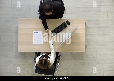 Asian Human Resource Manager in black suit shake hands to congratulate young candidate after the interview. Computer monitor, keyboard, mouse and resume are on wooden table. Top View Stock Photo