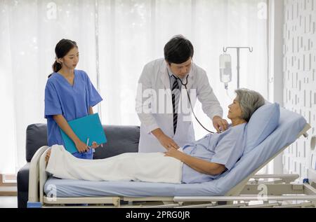 Senior female patient and doctor. Asian doctor use a stethoscope to check the heartbeat of the elderly patient. Young nurse stand next to the bed, holding document folder. Stock Photo