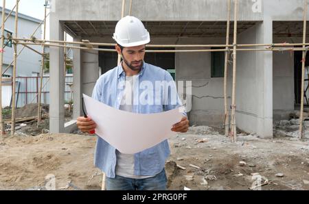 Young engineer in a construction helmet looking at real estate projects floor plan. The construction site has scaffolding made of wood. Stock Photo