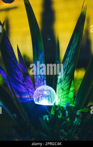 Night View Of Flowerbed Illuminated By Energy-Saving Solar Powered Colorful Multi-colored Lantern On Yard. Beautiful Small Garden With Blue Light, Lamp In Flower Bed. Garden Design Stock Photo