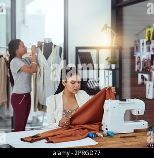 Checking her fabric. a young fashion designer sewing garments while a colleague works on a mannequin in the background. Stock Photo