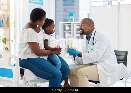 Doctor or pediatrician gives a teddy bear to a little girl after a medical appointment in a clinic with her mother. Small, happy child visiting the specialist for checkup in a healthcare hospital Stock Photo