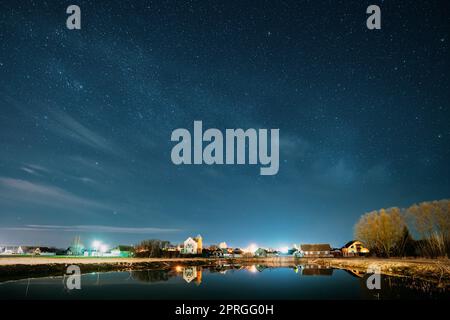 Belarus, Eastern Europe. Night Sky Stars Above Countryside Landscape With Lake Coast And Small Town Or Village. Natural Starry Sky Above Pond And Houses In Early Spring Night Stock Photo