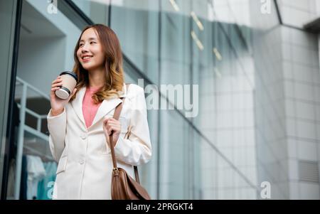 Happy business woman smiling hold paper cup of hot drink outdoor walking on street, Asian businesswoman holding coffee cup takeaway going to work she Stock Photo