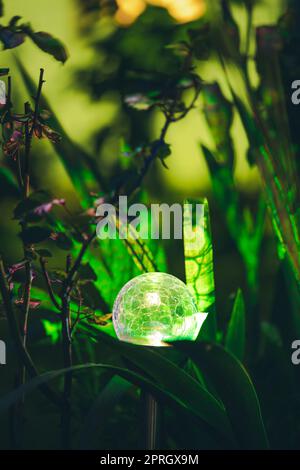 Night View Of Flowerbed Illuminated By Energy-Saving Solar Powered Colorful Multi-colored Lantern On Yard. Beautiful Small Garden With Green Light, Lamp In Flower Bed. Garden Design Stock Photo