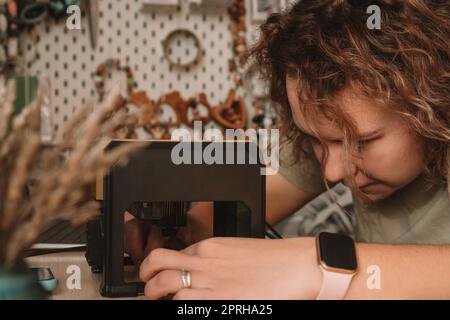 Curly haired woman holds item in desktop laser wood burning machine to inscribe name on surface of childrens wooden toy. Home production of teethers a Stock Photo