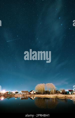 Belarus, Eastern Europe. Night Sky Stars Above Countryside Landscape With Lake Coast And Small Town Or Village. Natural Starry Sky Above Pond And Houses In Early Spring Night Stock Photo