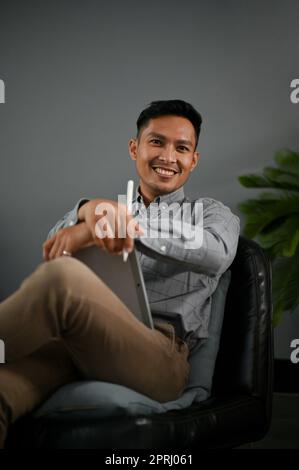 Handsome and smiling millennial Asian man in casual clothes sits on a black leather chair with his digital tablet. Stock Photo