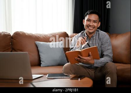 Smiling millennial Asian man making list or taking notes on his notebook while sitting on sofa in his living room. Stock Photo