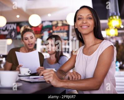 Who needs office hours. Portrait of a young woman sitting in a restaurant with two of her colleagues in the background Stock Photo