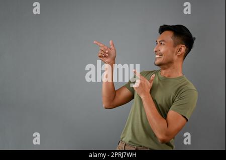 Smiling millennial Asian man in casual clothes pointing his finger aside on an empty space, recommending or showing choices, isolated on a grey backgr Stock Photo