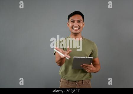 Smiling millennial Asian man in casual clothes with his digital tablet stands isolated on a grey studio background with a presenting gesture. Stock Photo