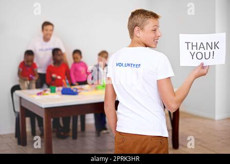 Everything helps. a volunteer holding a volunteers needed sign with volunteers working with little children in the background. Stock Photo