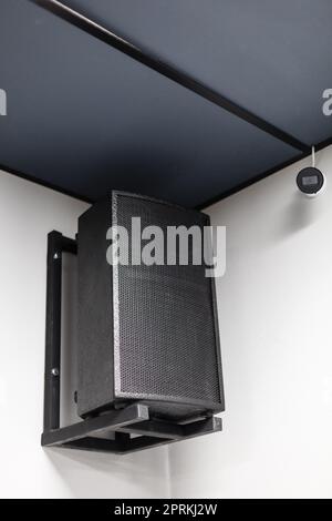 Close up of black big musical powerful speaker with protective grill hanging on wall in public place. Music speaker hangs on white wall near CCTV came Stock Photo