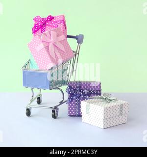 Small gift boxes of different colors with ribbons lies in shopping cart on a violet and blue pastel background. Stock Photo