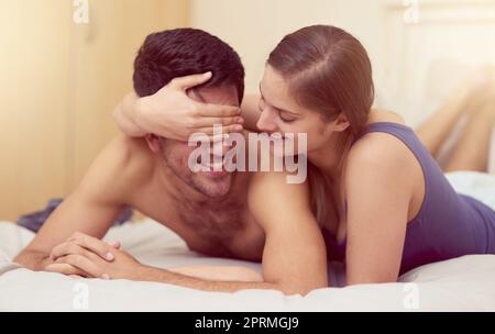 Guess who...a young woman playfully covering her boyfriends eyes while they lie in bed together. Stock Photo