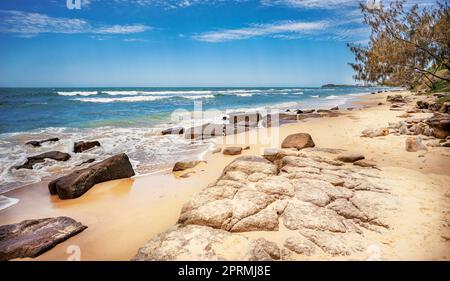 Ebb tide coming in on a rocky seashore. Surf waves break over shallow reef. Cloudy blue sky on a spring day on Sunshine Coast, Queensland. Stock Photo