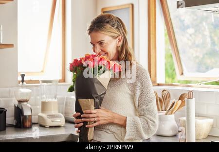 Flowers, smile, and elderly woman smelling rose in a kitchen, surprised by sweet gesture and or secret gift. Happy, romantic and kind surprise for mat Stock Photo