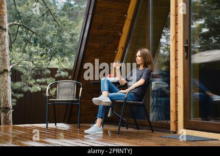 Cute woman sits on terrace of her forest house and drinks juice Stock Photo