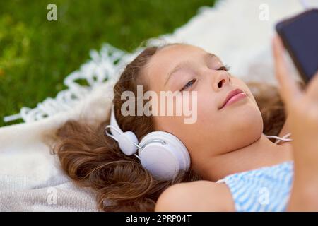 Chilling with some tunes. High angle shot of a young girl listening to music on her cellphone while lying on a blanket outside. Stock Photo