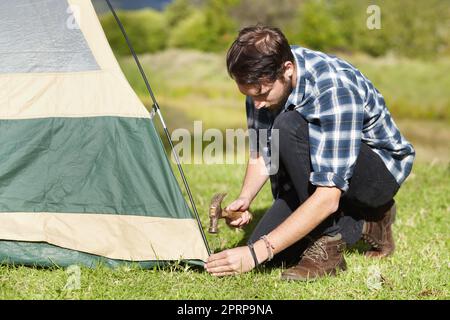 Better make sure its securely fastened. Handsome young man hammering a tent peg into the ground Stock Photo