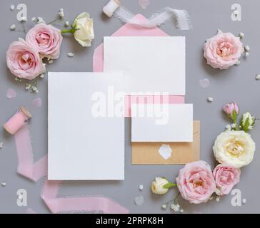 Blank paper cards and envelopes between pink roses on grey top view, wedding mockup Stock Photo