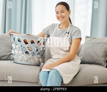 Cleaning, laundry and happy asian woman with on the sofa with a basket about to do washing of fold clothes in the living room at home. Portrait face housewife or maid doing chores and housework Stock Photo