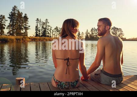 Enjoying the last rays of a summers day. an affectionate young couple in swimsuits sitting on a dock at sunset. Stock Photo