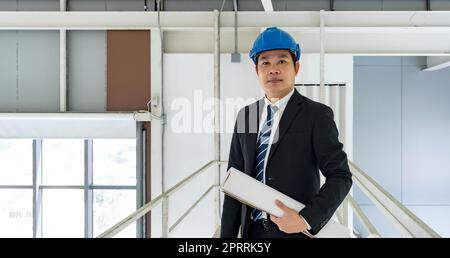 Asian businessman in black suit wearing a blue construction hat,  holding construction drawing. Inspector inspect the orderliness of the factory on the walkway. Stock Photo