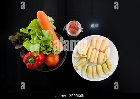 A glass bowl with a variety of fruits and vegetables is placed on the kitchen counter. Bell pepper, tomato, carrot and lettuce are in glass bowl. A dish with sliced apple was placed next to it. Stock Photo