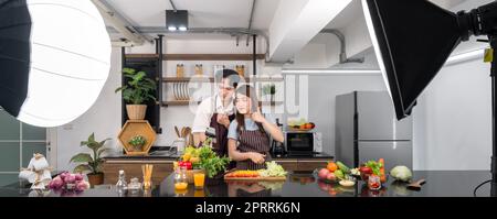 Asian couple spend time together in the kitchen. Young woman in apron cooking salad dish while his boyfriend recording vlog video for social blogger. Photography studio lights set up in the kitchen. Stock Photo