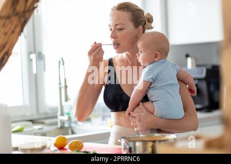 Woman cooking and tasing food while holding four months old baby boy in her hands Stock Photo