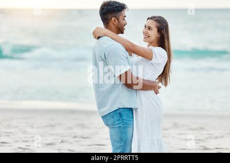 Couple love at beach, look in eyes and hug, with sunset over ocean in nature background or scene. Happy young, man and woman on sea sand smile together, sun setting over waves in backdrop or horizon. Stock Photo