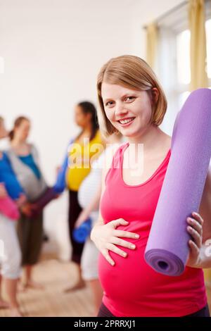 Glowing, healthy pregnancy. A young pregnant woman standing in a gym holding an exercise mat with a group of women in the background. Stock Photo