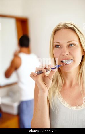 Getting ready together. A woman brushing her teeth while her boyfriend is getting ready in the background. Stock Photo