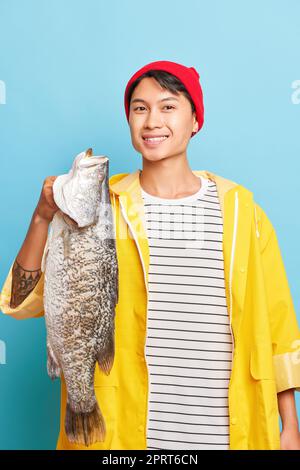 Smiling young fisherman holds big fish in his hand, wearing red cap yellow jacket, looks at camera with smile, good luck concept, copy space, high qua Stock Photo