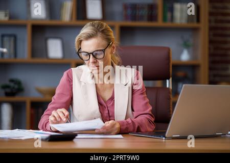 Portrait of adult businesswoman wearing eyeglasses working at office. Female manager doing paperwork, analyzing financial report, holding documents Stock Photo