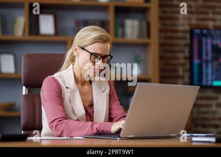 Portrait of adult businesswoman working with laptop at office. Female trader texting messages, doing paperwork with stock market graphs on background Stock Photo