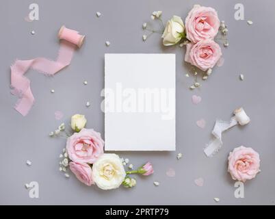 Blank paper card between pink roses and pink silk ribbons on grey top view, wedding mockup Stock Photo